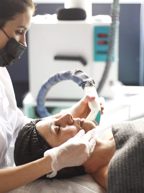 Female beautician in mask doing laser therapy procedure on face of relaxed woman during work in cosmetology clinic